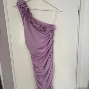 Lila oneshoulder  - Lång med oneshoulder och volangdetalj längs axeln. Klänningen har draperad effekt på sidan och är figurnära. För fler bilder kontakta mig. 