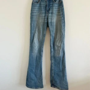 Blå bootcut jeans med hög midja - Snygga blå jeans med bootcut-ben och hög midja. Klassisk femficksmodell med dragkedja och knapp framtill. Jeansen har en lätt tvättad look och är tillverkade i ett robust denimtyg. Perfekta för dig som gillar en retrovibe och vill ha ett par jeans som sticker ut.