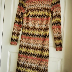 Mönstrad knälångklänning Pernilla Wahlgren - Färgstark klänning från Pernilla Wahlgren med zigzag-mönster i guld, rosa, brun, beige och svart. Klänningen har lång ärm och är stickad i ett glittrigt material som verkligen sticker ut. Perfekt för dig som vill ha något unikt och trendigt.