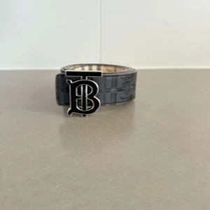 Svart Burberry bälte med TB-spänne - Snyggt svart bälte från Burberry med klassiskt rutmönster och präglad logga på remmen. Stort metallspänne med TB-monogram i silverfärg ger en lyxig vibe. Bältet är gjort i skinn och har en bred, stilren design som sticker ut.