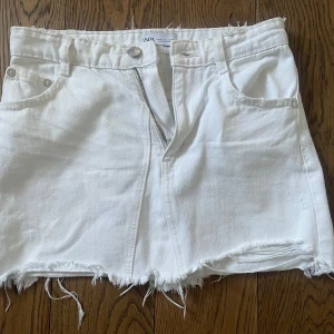 Vit jeanskjol med fransad kant från Zara - Vit kort jeanskjol från Zara med rå fransad kant och klassiska fem fickor. Kjolen har dragkedja och knapp framtill samt bälteshällor. Perfekt för en cool och avslappnad look.