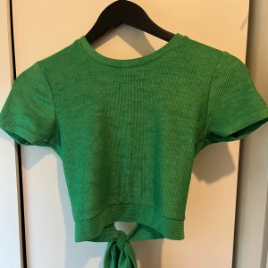 Grön ribbad croppad topp med knyt - Snygg grön croppad topp med kort ärm och ribbad struktur. Toppen har en cool öppen rygg och knytning. Perfekt för dig som vill sticka ut med en färgstark och trendig stil.