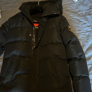 Svart pufferjacka från Zavetti Canada - Säljer en svart pufferjacka från Zavetti Canada med huva och snyggt broderat märke på ärmen. Jackan har dragkedja och knappar framtill samt två stora fickor. Perfekt för kalla dagar och riktigt stilren look.