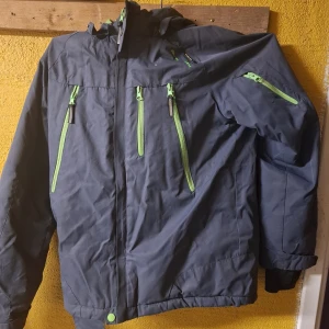 Grå vinterjacka On The Peak 164 - Fakta: Grå vinterjacka från On The Peak med limegröna dragkedjor och detaljer. Jackan har huva med grön insida, flera fickor med dragkedja och justerbara ärmslut. Perfekt för kalla dagar och outdoor-aktiviteter. Bra för: Barn som behöveroömma kläder. OBS: Se sista bilden. Liten skada. 