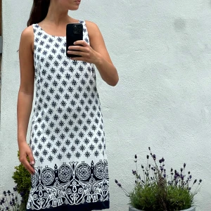 Vit och marinblå mönstrad sommarklänning - Snygg ärmlös kortklänning i vitt med marinblått grafiskt mönster och dekorativ bård längst ner. Klänningen har rak passform och rund halsringning, perfekt för varma dagar. Materialet känns lätt och luftigt, vilket gör den superbekväm att bära.