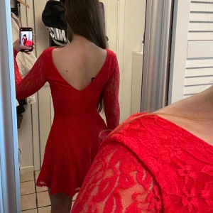 Röd spetsklänning med öppen rygg - Supersnygg röd kort klänning i spets med långärmade ärmar och djup v-ringad rygg. Klänningen har en figurnära passform upptill och utsvängd kjol. Perfekt för dig som vill sticka ut med en feminin och trendig look.