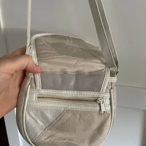 Beige axelväska i skinn med patchwork - Snygg beige axelväska i skinn med patchwork-design och justerbar rem. Väskan har ett lock med tryckknapp, innerfack och en dragkedjeficka på baksidan. Perfekt storlek för det viktigaste och stilren look.