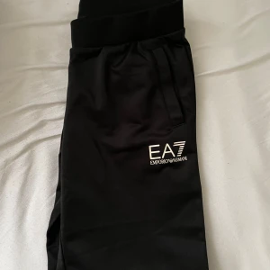 Svarta EA7 Emporio Armani mjukisbyxor - Svarta mjukisbyxor från EA7 Emporio Armani med vit logga på benet. Byxorna har elastisk midja och raka ben, tillverkade i mjukt material som känns skönt mot huden. Perfekta för chill eller träning. (12-13 år) köpt för 699kr! Förra året 