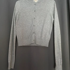 Grå kofta från Gina Tricot - En stilren grå kofta från Gina Tricot med rund halsringning och knappar framtill. Koftan är långärmad och har en klassisk, enkel design som passar till det mesta. Tillverkad i mjukt material som känns skönt mot huden.