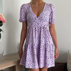 Lila sommarklänning - Supersöt kortklänning i ljuslila med små blommor, v-ringad omlottmodell och puffiga kortärmar. Klänningen har volangdetaljer och knyt i sidan.