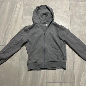 Grå hoodie från Polo Ralph Lauren - Snygg grå hoodie från Polo Ralph Lauren med dragkedja och klassisk vit broderad logga på bröstet. Tröjan har huva, kängurufickor och är tillverkad i mjuk bomullsmix. Perfekt för en chill och stilren look.