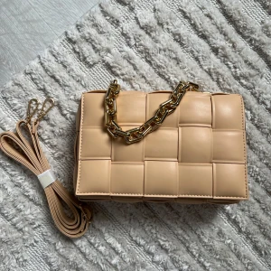 Beige quiltad axelväska med kedja - Säljer en beige axelväska i quiltad design med breda flätade rutor. Väskan har en chunky guldfärgad kedja som handtag och en avtagbar axelrem i matchande beige. Stängs med magnetknapp och har svart innerfoder. Perfekt för dig som gillar trendiga accessoarer.