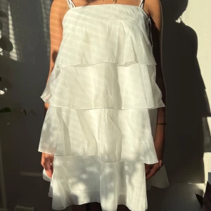 Vit volangklänning med tunna axelband - Superfin vit kort klänning med flera lager volanger och tunna axelband. Klänningen har en luftig och flowy känsla, perfekt för varma dagar. Materialet är lätt och ger en snygg siluett. Den är från Ginatricot, aldrig använt utan endast tagit av prislappen på. Volangerna är i hop sytt lite då jag inte ville ha den för puffig men inget så stor skillnad. Den sträcker sig precis innan knäna på mig som är 162cm💕💕🫶🏼🫶🏼