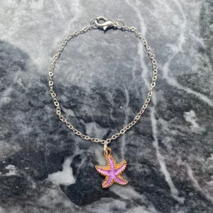 Armband med lila sjöstjärna - Snyggt armband med tunn silverfärgad kedja och en lila sjöstjärneberlock med guldkant. Perfekt accessoar för dig som gillar havsinspirerade smycken och vill ha något unikt och lekfullt på handleden.
