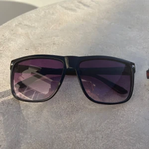 Svarta solglasögon med lila tonade glas - Snygga svarta solglasögon med bred båge och lila tonade glas. Klassisk fyrkantig form med små silvriga nitar i hörnen för en extra detalj. Perfekta för soliga dagar och ger en cool vibe till din outfit.