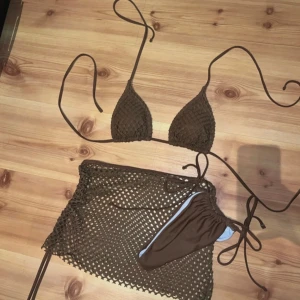 Brun virkad bikini med kjol - Snygg brun bikini med trekantsöverdel och knyt i sidorna. Setet inkluderar en matchande virkad kjol som ger en trendig strandlook. Bikinin har en enkel, stilren design och kjolen är genombruten för extra sommarkänsla.