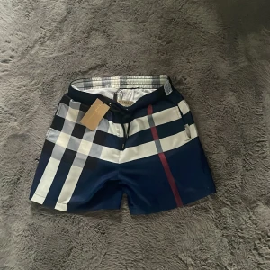Rutig Burberry badshorts blå/vit - Snygga badshorts från Burberry med klassiskt rutigt mönster i blått, vitt och svart samt en röd detalj. Shortsen har snörning i midjan och meshfoder på insidan. Perfekta för stranden eller poolen, med en stilren och exklusiv känsla. Storlek M med sitter som S