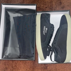 Svarta Saint Laurent sneakers - Snygga svarta sneakers från Saint Laurent med vit broderad logga på sidan. Skorna har en vit platt sula och är tillverkade i canvas och skinn. Klassisk snörning och stilren design som passar till allt. Kommer i originalkartong.