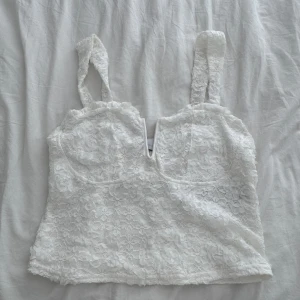 Vit spetsig korsett-topp - Superfin vit korsett-topp med breda axelband och v-ringning. Toppens yta är täckt av blommig spets som ger en romantisk och trendig vibe. Perfekt att styla med jeans eller kjol för en cool look.