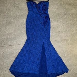 Blå långklänning med pärldetaljer - Snygg blå långklänning med figurnära passform och utsvängd nederdel. Klänningen är axelbandslös och har vackra broderier och pärlor upptill. Materialet är spetsliknande med struktur och en slits framtill för extra effekt. Perfekt för bal eller fest. Jag har sytt till axelband men de är enkel att ta bort om så önskas. Lite pärlor som hänger men det går att fixa enkelt. På lappen står det att det är en storlek 40 men personligen hade jag sagt att den passar mer för en 38.💗