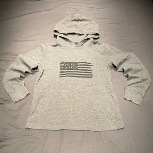 Grå hoodie från Polo Ralph Lauren - Säljer en grå stickad hoodie från Polo Ralph Lauren med tryck av en flagga och märkesnamnet på bröstet. Tröjan har långa ärmar och huva, perfekt för en avslappnad stil. Den är liten i storleken