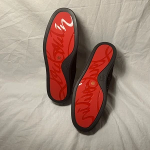 Christian Louboutin svarta sneakers med nitar - Svarta sneakers från Christian Louboutin i mocka med ikonisk röd sula och svarta nitar på tån. Skorna har snörning och rund tå, samt lyxig känsla med guldfärgad logga på innersulan. Perfekt för dig som vill sticka ut med edgy detaljer.