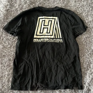 Svart Hollister t-shirt - Svart t-shirt från Hollister i storlek XXL med stort grafiskt tryck och logga på ryggen samt mindre logga framtill. Tillverkad i 100% bomull, klassisk passform och rund hals. Perfekt för dig som gillar streetwear och tydliga prints.