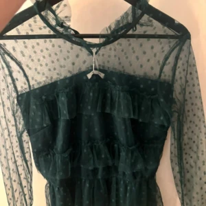 Mörkgrön meshblus med volanger - Supersnygg mörkgrön blus i mesh med prickigt mönster och flera lager volanger framtill. Blusen har långa, transparenta ärmar och hög hals. Perfekt för dig som vill sticka ut med en cool och trendig look. Nypris 500kr
