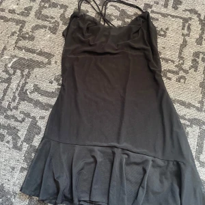 Svart kort klänning med volang - Svart kort klänning med tunna axelband som korsas i ryggen. Klänningen har en lätt volang nertill och är sydd i ett tunt, något transparent tyg. Snygg och enkel modell som ger en stilren look.