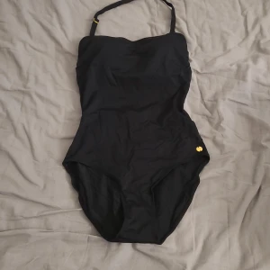Svart baddräkt från Twilfit S - Snygg svart baddräkt från Twilfit med vadderade kupor och justerbara axelband med gulddetaljer. Klassisk och stilren modell som sitter tight och ger bra stöd. Perfekt för strand eller poolhäng i sommar. Endast använd 1 gång. 