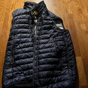 Svart dunväst från Parajumpers - Snygg svart dunväst från Parajumpers med quiltad design och hög krage. Västen har två dragkedjeförsedda fickor framtill och coola detaljer som märkets logga på insidan. Perfekt för lager-på-lager och funkar året runt.