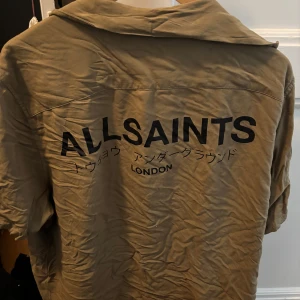Beige skjorta från AllSaints XL - Snygg grön/beige skjorta från AllSaints i relaxed fit med svart tryck på både bröstet och ryggen. Skjortan har klassisk krage, korta ärmar och knäppning framtill. Perfekt för dig som gillar streetwear och vill sticka ut med en cool logga.
