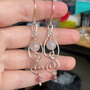  Silverörhängen med stenar av rosenkvarts  - Unika örhängen i handformad silverfärgad tråd med spiralformade detaljer. De har droppformad design och är dekorerade med runda pärlor i ljusrosa och lila toner. Perfekta för dig som gillar boho eller artsy stil och vill sticka ut med något personligt. Går att anpassa efter egna önskemål på färger.