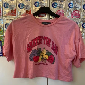 Rosa croppad topp från Primark XS - Croppad rosa topp från Primark med tryck av frukter och texten 'Squeeze the day'. T-shirten har korta ärmar och rund halsringning. Perfekt för dig som gillar färgglada och roliga prints. Gjord i mjuk bomull för en skön känsla.