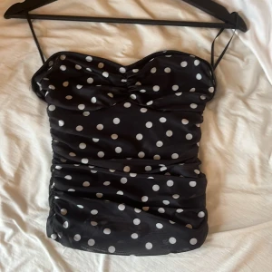 Svart prickig bandeau tubtopp - Supersöt svart bandeau tubtopp med vita prickar och rynkade detaljer längs sidorna. Toppen är axelbandslös och har en figurnära passform som ger en snygg siluett. Perfekt för dig som vill sticka ut med ett lekfullt mönster. Toppen passar alla då den är stretchig vid sidorna med ett tunt tyg. Pris 170kr plus frakt! OANVÄND