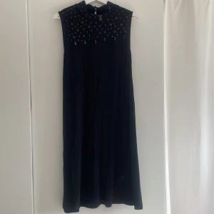 Svart långklänning med pärlor från SOYA - Svart ärmlös långklänning från SOYA med hög hals och dekorativa svarta pärlor och paljetter upptill. Klänningen har en rak, elegant siluett och knäppning i nacken. Perfekt för dig som vill sticka ut med en stilren look.