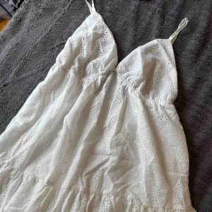 Vit somrig klänning med broderade blommor - Supersöt vit kortklänning med tunna axelband och v-ringning. Klänningen har broderade blommor över hela tyget och en lätt volang nertill. Perfekt för varma dagar och har en luftig känsla. Använd en gång