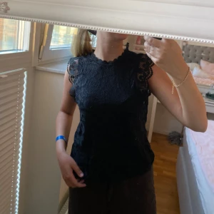 Svart spetsblus med volangärmar - Supersöt svart blus i spets med fina volangdetaljer på ärmarna och runt halsen. Blusen är ärmlös och har ett romantiskt mönster i spetsen som ger en elegant vibe. Perfekt att styla med jeans eller kjol för en trendig look.