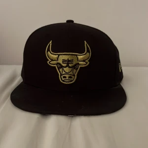 Svart Chicago Bulls keps från New Era - Snygg svart snapback-keps från New Era med Chicago Bulls-logga broderad i guld framtill och texten 'BULLS' bak. Klassisk platt skärm och justerbar passform. Perfekt för dig som vill ha en sportig och streetinspirerad stil.