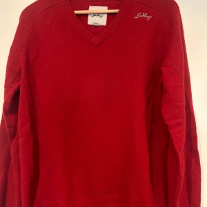 Röd ulltröja med v-ringning Lundhags - Röd stickad ulltröja från Lundhags med v-ringad hals och broderad logga på axeln. Tröjan har långa ärmar och ribbade muddar vid ärmslut och nederkant. Perfekt för dig som gillar klassisk stil med en färgstark touch. Stl M