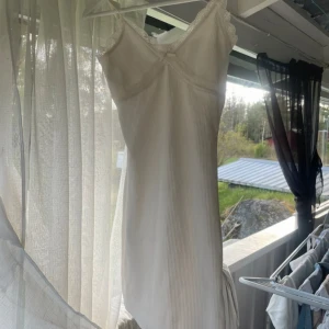 Vit ribbad kortklänning från H&M - Supersöt vit ribbad kortklänning från H&M i storlek XS. Klänningen har tunna axelband, spetsdetaljer längs urringningen och en liten rosett framtill. Perfekt för sommarens varma dagar och har en feminin, enkel look.