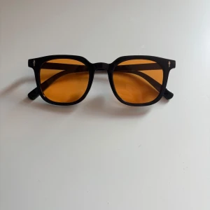 Svarta solglasögon med orange glas - Snygga solglasögon med svart, fyrkantig båge och coola orangefärgade glas. Klassisk modell med markerade kanter och diskreta silvriga detaljer vid sidorna. Perfekta för att sticka ut och ge en retrovibe till din stil.