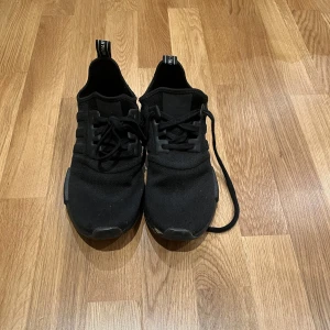 Svarta Adidas NMD sneakers - Säljer ett par helsvarta Adidas NMD sneakers med klassisk mesh-ovandel och svarta detaljer. Skorna har snörning, platt sula och diskret Adidas-logga på hälen. Perfekta för dig som gillar stilrena och bekväma sneakers.
