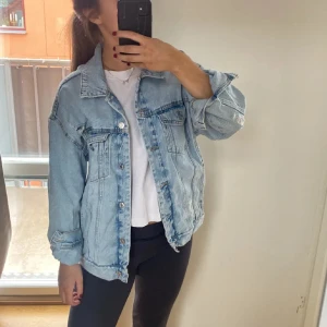 Oversized ljusblå jeansjacka från PULL&BEAR - Säljer en oversized ljusblå jeansjacka med klassiska bröstfickor och knappar framtill. Jackan har en avslappnad passform, slitna detaljer på axeln och är tillverkad i denim. Perfekt för dig som gillar en chill och trendig stil.