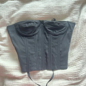 Svart korsett med snörning - Snygg svart korsett med formade kupor och smala axelband. Toppen har markerade sömmar framtill och snörning i ryggen med svarta satinband för en justerbar passform. Perfekt att styla med jeans eller kjol för en cool look.
