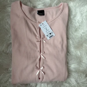 Ljusrosa ribbad topp med rosetter Gina Tricot - Supersöt ljusrosa ribbad topp från Gina Tricot med lång ärm och dekorativa rosetter framtill. Toppen har rund halsringning och är tillverkad i mjuk bomullsmix. Perfekt för dig som gillar gulliga detaljer.