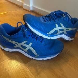 Blå Asics Gel-Phoenix 10 sneakers - Säljer ett par blå Asics Gel-Phoenix 10 sneakers med vita och ljusgula detaljer. Skorna har snörning, ventilerande mesh-ovandel och en vit sula med dämpning. Perfekta för löpning eller träning, snygg sportig look och bekväm passform.