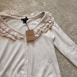 Vit kofta med volangkrage från H&M - Supersöt vit kofta från H&M med lång ärm och knäppning framtill. Koftan har en bred volangkrage som ger en gullig och trendig look. Perfekt att styla med jeans eller kjol för en extra touch. Materialet är mjukt och känns skönt mot huden. Varan är i bra skick och har lappen kvar från Sellpy! OBS! Den har en ljusrosa/vit färg, så inte helt beige