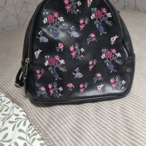 Svart ryggsäck med broderade blommor - Säljer en svart ryggsäck i skinnimitation med broderade blommor i rosa, lila och vitt. Väskan har dragkedja upptill och justerbara axelremmar. Perfekt för dig som vill ha en söt och trendig accessoar med plats för det viktigaste.