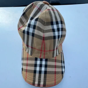 Beige rutig keps från Burberry - Helt oanvänd och snygg keps från Burberry i klassiskt beige rutigt mönster med svarta, vita och röda detaljer. Kepsen har böjd skärm och justerbar rem bak. Tillverkad i bomull med insida i svart tyg. Perfekt för dig som vill sticka ut med en ikonisk accessoar.